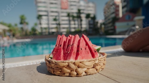 Close-up of a woven basket filled with fresh watermelon slices by an outdoor swimming pool on a sunny summer day. A woman in a blue swimsuit reaches for a slice, creating a relaxed vacation atmosphere