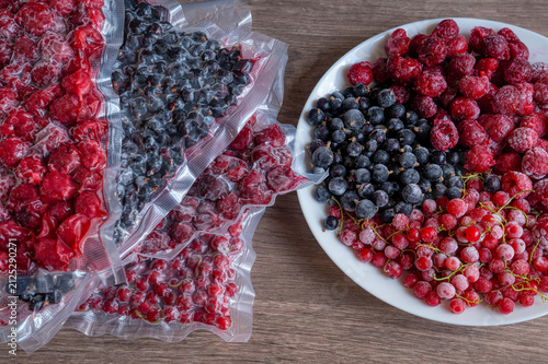 Plate with frozen berries beside stack of vacuum sealed plastic bags with raspberries black currants cherries red currants on wooden table