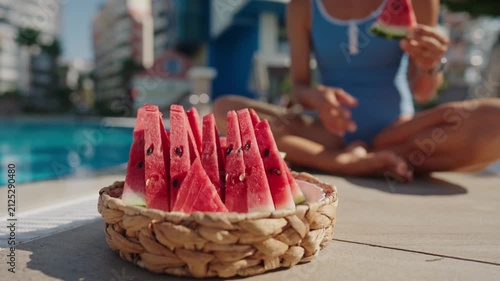 Close-up of a woven basket filled with fresh watermelon slices by an outdoor swimming pool on a sunny summer day. A woman in a blue swimsuit reaches for a slice, creating a relaxed vacation atmosphere