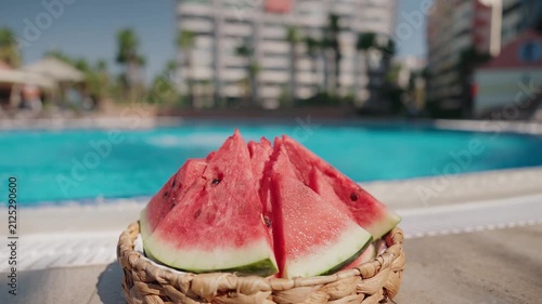 Close-up of a woven basket filled with fresh watermelon slices beside an outdoor swimming pool on a bright summer day. In the blurred background, a young boy jumps into the pool, adding a sense of fun