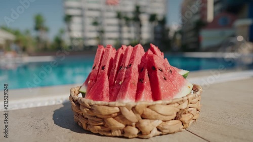 A basket filled with fresh slices of ripe watermelon sits by a sparkling outdoor swimming pool on a bright summer day. The vibrant red fruit contrasts beautifully with the crystal-clear blue water