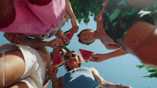 Low-angle view of a family standing in a circle outdoors on a sunny summer day, holding fresh watermelon slices and enjoying a healthy snack together. The camera looks up toward the blue sky, creating