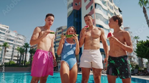 A happy family enjoys eating fresh watermelon together by an outdoor swimming pool on a sunny summer day. Parents and children share juicy slices of seasonal fruit during a relaxing resort vacation