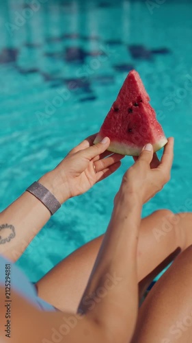 A woman relaxes by an outdoor swimming pool holding a fresh slice of watermelon on a sunny summer day. Viewed from behind, she enjoys a peaceful vacation moment while cooling her feet in the water
