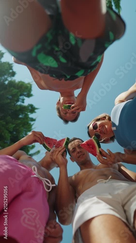 Low-angle view of a family standing in a circle outdoors on a sunny summer day, holding fresh watermelon slices and enjoying a healthy snack together. The camera looks up toward the blue sky, creating