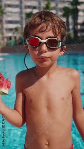 A smiling boy wearing swimming goggles enjoys a fresh slice of watermelon by an outdoor swimming pool on a bright summer day. The child stands poolside after swimming, eating juicy seasonal fruit
