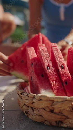 Close-up of a woven basket filled with fresh watermelon slices by an outdoor swimming pool on a sunny summer day. A woman in a blue swimsuit reaches for a slice, creating a relaxed vacation atmosphere