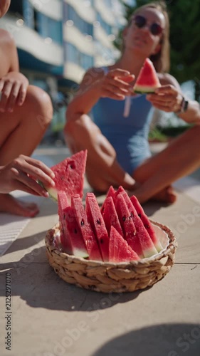 Close-up of a woven basket filled with fresh watermelon slices by an outdoor swimming pool on a sunny summer day. A woman in a blue swimsuit reaches for a slice, creating a relaxed vacation atmosphere