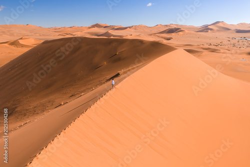 Aerial view shows a lone hiker walking a sharp sand dune ridge in Sossusvlei near Deadvlei, Namibia, with apricot dunes and dark wind shadow in bright daylight.