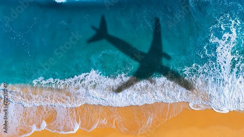Aerial view of a passenger airplane shadow falling onto the turquoise blue ocean water waves and sandy tropical beach during a summer flight