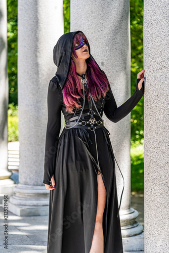 Young woman wearing a black gothic dress, decorative mask and hood poses beside granite columns in a neoclassical pavilion, creating a mysterious alternative fashion portrait outdoors