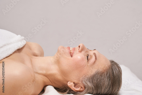 Elderly Woman Lying Relaxed On Spa Bed With Smiling Face And Towel In Soft Neutral Treatment Room.