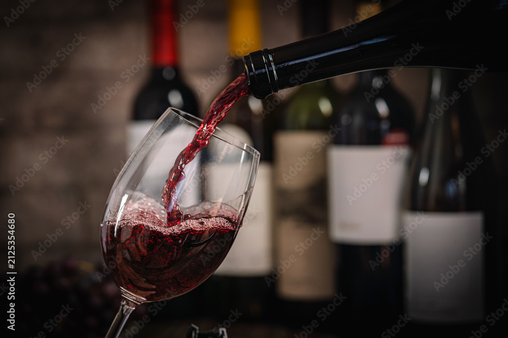 Obraz premium A dark, elegant still life of red wine being poured into a glass with wine bottles and fresh grapes in the background. Perfect for wine-themed compositions