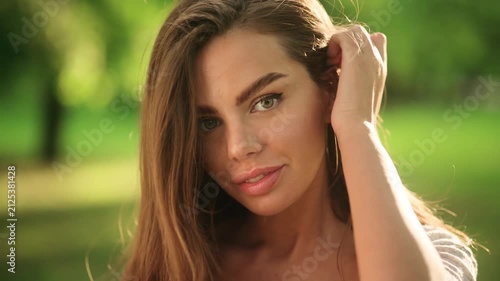 Close up portrait of a beautiful attractive young woman looking at camera and touching her hair in park. Pretty female enjoying warm sunny weather on blurred nature background