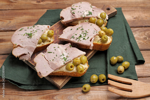 Sliced rustic bread topped with savory ham, creamy hummus spread, green pitted olives, and fresh chopped parsley served on a wooden board over a dark green napkin.