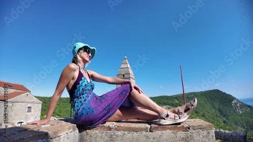 Woman in a colorful dress and hat relaxing on a stone wall. Enjoying the panoramic view of the historic Lubenice village. A medieval settlement on Cres Island. Croatia of Cres Island in Croatia