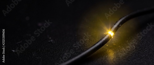 Glowing yellow electrical spark jumping from damaged black power cable isolated against dark background showing wiring danger.