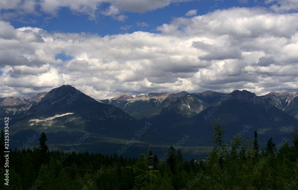 Obraz premium A detailed panoramic view focusing on rugged, dark mountain peaks under a heavily textured blanket of white and grey cumulus clouds with a deep pine forest in the foreground.