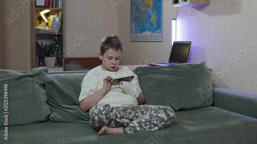 Boy with cast on arm sitting on sofa and looking at smartphone screen in home setting. Rest during home rehabilitation after injury. Remote learning during outpatient treatment with injured limb.