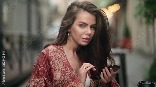 Beautiful attractive young woman texting on smartphone at outdoor cafe table, then looking at camera. Pretty female with coffee cup sitting on blurred urban street background