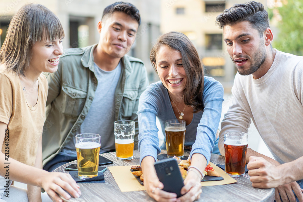Obraz premium Friends smiling at a smartphone while sharing beers at an outdoor cafe. Multiethnic millennials and gen z gather around the table for a digital memory. Social media concept