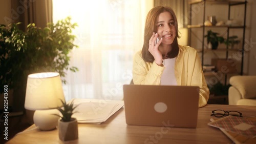 Beautiful attractive young woman sitting at desk with laptop and talking on phone at home office. Happy pretty female student or freelancer answering call in a cozy sunlit room