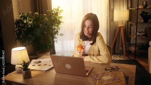 Beautiful attractive young woman having a video call on laptop and peeling a mandarin. Happy pretty female student or freelancer conversing online at home office with sunny window