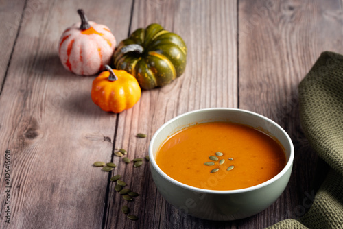 owl of warm pumpkin soup sprinkled with seeds, surrounded by mini pumpkins and green towel on wooden surface