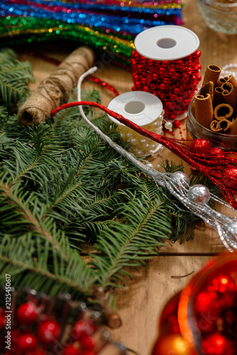 Vertical closeup of christmas crafting materials on wooden table with pine branches glitter ribbons red beads cinnamon sticks and twine for cozy holiday DIY decoration projects
