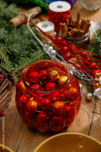 Vertical still life of red glass jar filled with shiny red and gold christmas baubles surrounded by pine branches cinnamon sticks ribbons and glitter garlands on wooden table