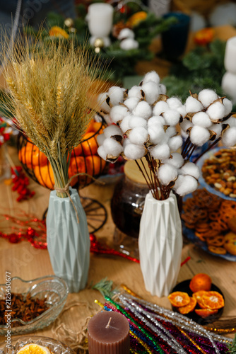 Vertical still life of dried wheat and cotton branches in geometric vases surrounded by tangerines candles glitter wires and spices on wooden table for cozy holiday decor