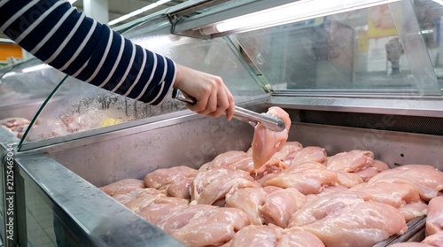 Hand in a striped sleeve using steel tongs to pick up a piece of fresh raw chicken breast from a chilled metal display tray in a retail food store