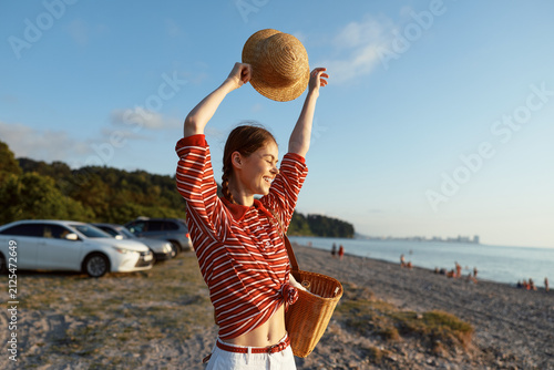 Foto A smiling woman in a striped shirt lifts a hat above her head on a sunny beach, holding a wicker basket