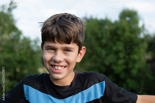 Smiling young boy outdoor portrait wearing blue and black shirt with casual happy expression. The guy has nice smile, but his teeth are crooked, they are growing crookedly, he needs braces