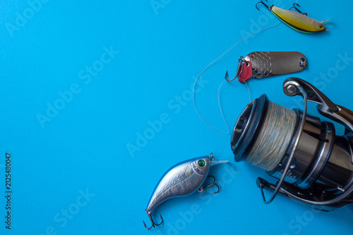 Quadro su tela Close‑up of a metallic fishing reel and three colorful lures on a bright blue background