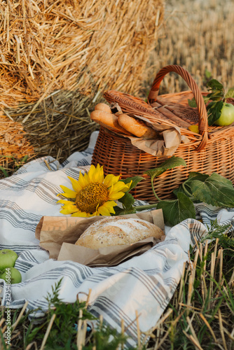 A woven basket filled with bread and apples sits beside a golden hay bale, with a sunflower and rustic blanket completing the countryside scene.