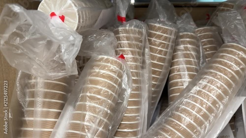 Stacks of disposable plastic drinking cups are packed in transparent bags and cardboard boxes, neatly arranged for sale in a large retail store for parties, events and everyday beverage service.
