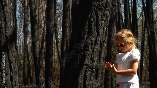 cheerful girl sunglasses white T-shirt stands burnt tree forest, touching hand, examining soot marks fingers pointing camera. It's sunny, bright spring day. consequences of a forest fire