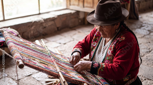 An elderly indigenous Andean woman wearing a traditional hat and embroidered jacket works on a colorful textile using a manual loom in Peru.