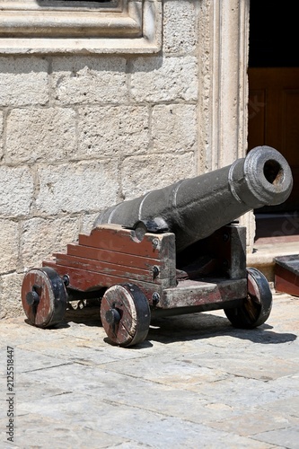 Canvastavla Close-up of an ancient black cast-iron cannon mounted on a brown wooden carriage with wheels, standing outdoors against a vintage stone fortress wall
