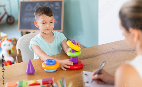 Boy sits at table and builds pyramid out of plastic toy cups. Childs behavior as marker of mental and mental health and development. Developing and maintaining skills for children with special needs