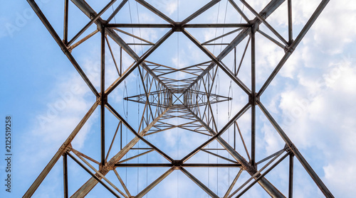 A low-angle, symmetric view looking up through the metal framework of a high-voltage electrical pylon against a bright blue sky with clouds.