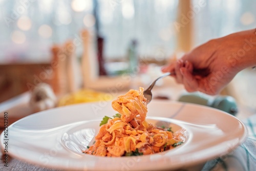 Фотография Hand twirls fresh orange cream pasta on a silver fork above a ceramic plate with