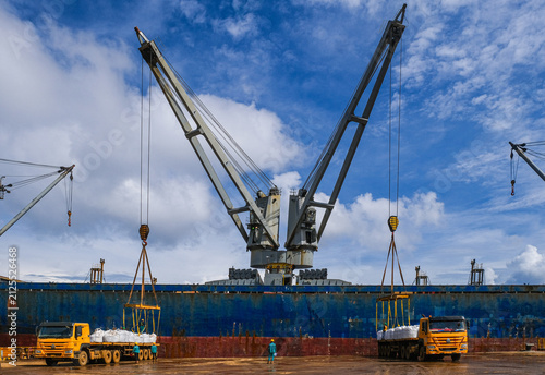 Rear view of a dock worker standing in front of a cargo ship between two trucks supervising the loading and unloading of merchandise, Indonesia