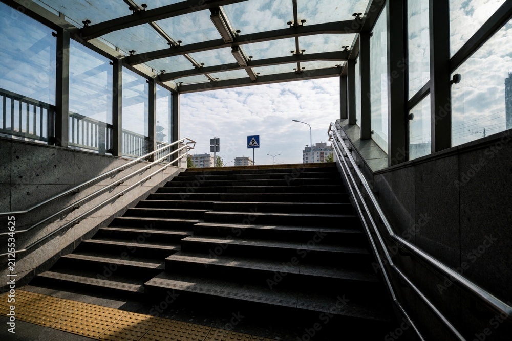 Fototapeta premium Concrete stairs lead from underground subway exit to city street level. Glass roof covers pedestrian passage staircase with metal railings. Sunny day view includes outdoor crosswalk sign, modern