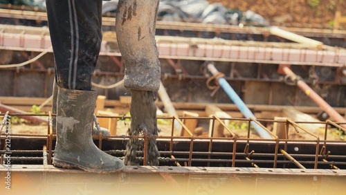 Worker wearing protective rubber boots standing on formwork while pouring fresh concrete mix from a pump hose into a building foundation structure with rebar