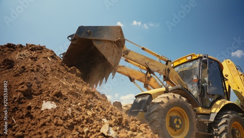 Принти на полотні Yellow backhoe loader moving and scooping brown dirt, performing groundworks at