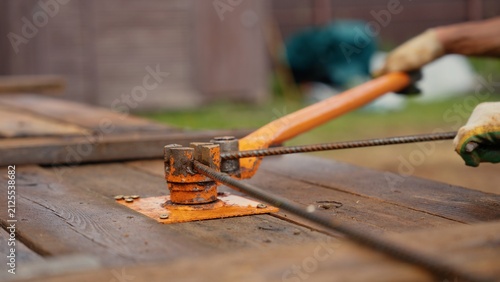 Worker's gloved hands operating a manual rebar bender, shaping steel reinforcement bars on a wooden surface for a construction project, emphasizing precision and physical labor in building
