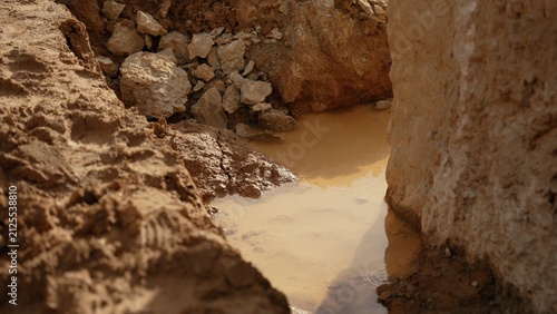 Deep excavation trench with muddy water collecting at the bottom, showing exposed earth layers, rocks, and wet soil on an active construction worksite