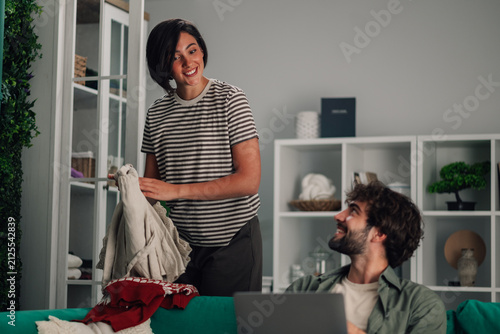 Foto Happy couple interacting at home, woman folding laundry while man works on lapto